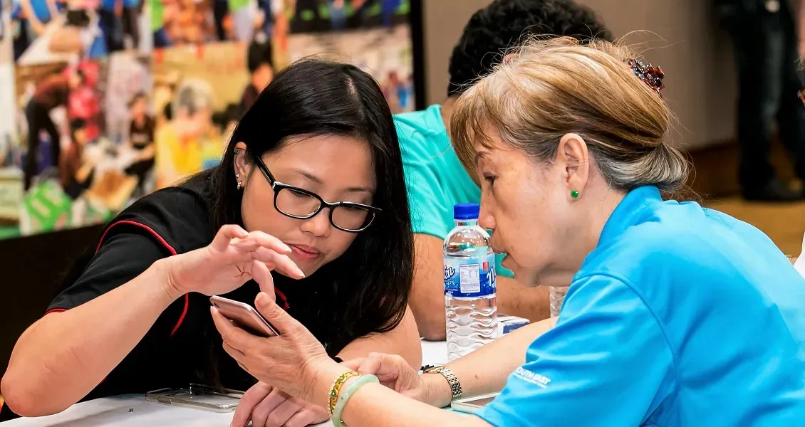 Two people are seated at a table, one demonstrating a mobile phone to the other with a colorful background behind them.